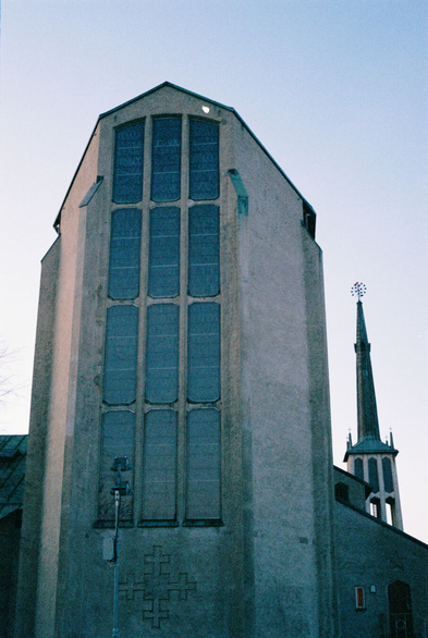 Photo of the back (or East) side of a large church built in bare concrete. The tall and narrow wall on the East end has twelve tall and narrow stained-glass windows, but the art on the windows can't be made out in the dim light. Instead, the windows reflect the clear sky in a gradient from light blueish-grey at the bottom to dark blue at the top. Slightly to the side in the background, a free-standing bell tower with an open construction and a very tall and thin copper spire can be seen.
