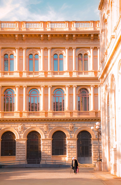 This photograph is of Piazza Minghetti and the grand facade of a classical, multi-story building (which is the post office). It is bathed in a warm, golden sunlight. The architecture has rows of arched windows with decorative columns on either side. There is also an ornate balustrade along the roofline, and large entry points on the bottom. In the foreground there is a  single person wearing a dark coat and carrying a pink bag who is walkign across the paved courtyard. There is a street lamp which stands nearby on the right, casting a shadow against the base of another building (which is a bank) that frames the edge of photo.