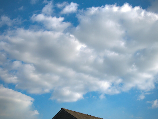 A large fluffy, puffy white cloud hangs in a bright blue sky on a sunny day. At the bottom of the image a small sliver of the peak of a slate roof peeks into the frame.