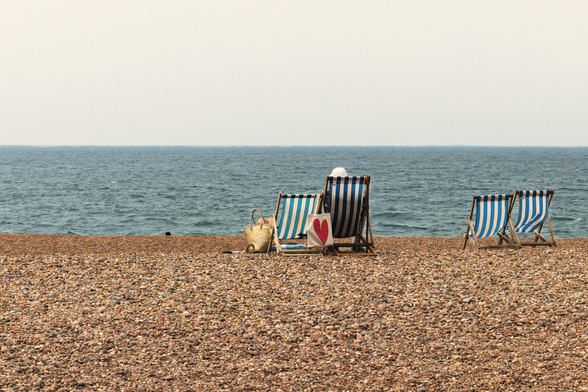 A wide pebble beach stretches across the foreground, leading to a calm blue-grey sea beneath a pale, hazy sky. Near the centre, two traditional blue-and-white striped deckchairs face the horizon. One is occupied by a person in a white sunhat, while another person reclines beside them, mostly hidden from view. Between the chairs sits a cream tote bag with a large red heart, alongside a woven straw beach bag. To the right, another pair of empty deckchairs stands alone, separated by open space.