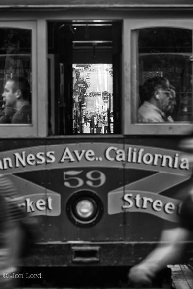 This is a black and white street photo in portrait format of the rear of a San Francisco cable car with blurred people walking in the foreground and the street beyond view through the car's centre window. California Street, San Francisco (2012).

In the foreground are the motion blurred arms of those pedestrians crossing the street behind the stationary cable car. The cable car is viewed from behind, at a distance of a few metres and filling the the entire image. Painted on the back of the car are the words 'Van Ness Ave.. California St' and below 'Market Street' with the number 59 and a single headlight in the centre. Above are three vertical rectangular windows, the two outer windows have passengers sitting facing outwards and viewed in profile with just their heads and shoulders in view. Through the centre window is a view of California Street beyond. Initially the street is flat with numerous people crossing the road and traffic driving along it. After about one hundred metres the road climbs upwards steeply for about a hundred metres, levels off for a short distance before climbing steeply again for a further hundred metres. This being repeated three times before disappearing out of view at the top of the photo. Visible in the centre of the road and climbing the hill are the two parallel sets of rail tracks (and centre groove) that the cable cars run along. 