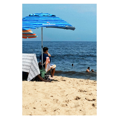 A boy sits under a beach umbrella with dark and light blue stripes on a sandy shore, licking an ice cream cone and looking out to sea. Tiny beachgoers poke their heads above the water. Another umbrella, with blue and orange stripes, pushes in from left frame. The sky is a clear blue with a hint of clouds.