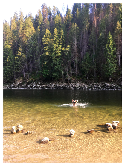 View of a wide stretch of river leading to a rocky bank and a hill covered with tall conifers on the far side. At the shallow end near camera are several rock piles previously made by visitors, surrounded by sunlit ripples. Further off, a swimmer emerges from the deeper water, hands smoothing back her hair, sending up a sunlit spray of droplets that contrasts with the darker reflection of the trees.