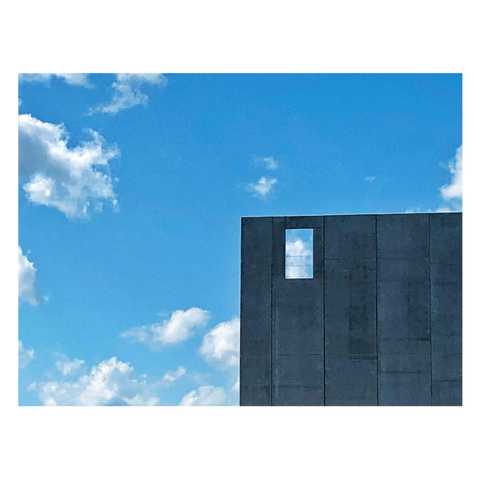 View of a blue summer sky with scattered puffy white clouds. The lower right quadrant of the image is occupied by a gray building facade of unpainted vertical panels. A single empty window frame at the facade’s upper left corner — really just a rectangular cutout — shows the sky and clouds behind, revealing that the structure is a flat freestanding surface, not a three-dimensional building. I find the image to be minimalist and evocative of one of Magritte’s sky paintings.