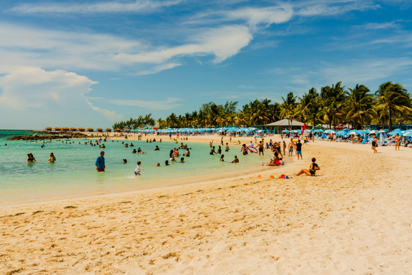 Tourists sit at blue umbrellas at the shore and float in the shallow blue-green waters at The Bahamas. A  forest of palm trees is behind them.