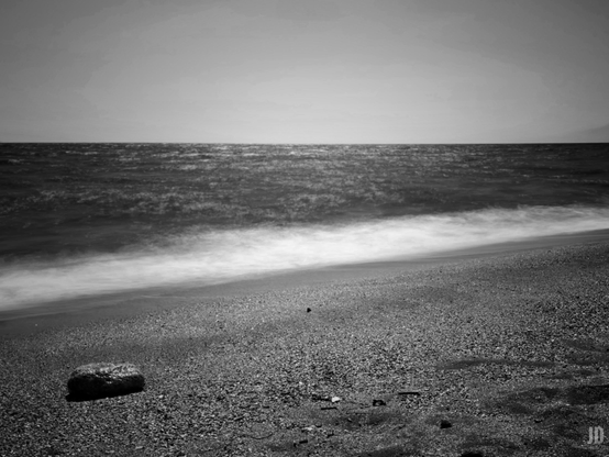La imagen muestra una playa en blanco y negro con una orilla de arena y pequeñas piedras. El mar aparece en movimiento, con las olas difuminadas, lo que sugiere una exposición larga. El horizonte separa el agua del cielo despejado y en primer plano se distingue una roca sobre la arena.