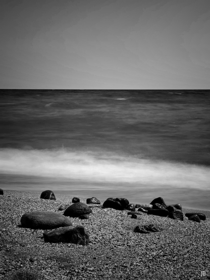 La imagen muestra una playa de guijarros en blanco y negro, con varias rocas dispersas en la orilla. El mar aparece con un efecto sedoso, como resultado de una exposición larga, y el horizonte separa claramente el agua del cielo despejado. La escena transmite calma y minimalismo.
