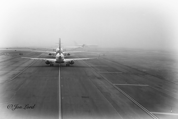 This is a black and white photo in landscape format of a busy airport taxiway surrounded by fog with a long line of aircraft waiting to takeoff. JFK, New York (2011).

One third of the way to the right of the left edge of the photo is a long solid line stretching along the centre of the taxiway and away from the camera. A short distance to the right of the centre line (about ten cm) is a line of lights inset into the tarmac, spaced every few metres. About ten metres to the left and right of the centre line are two parallel solid lines marking the edge of the taxiway, one either side, though the tarmac continues for a distance of another five or ten metres beyond. Further to the right is a wide grass area before a small portion of a parallel runway is in view. Surrounding this scene is a bright, white, morning fog with a visibility of no more than five hundred metres. In front of the camera is a row of five aircraft on the taxiway waiting for their turn to take off. The closest aircraft is about ten metres from the camera, is a Boeing 737 narrow body, shorthaul aircraft with two jet engines slung under the wings and small winglets on the wingtips. Beyond the queue of aircraft is a further larger aircraft, about five hundred metres away, at right angles to the taxiway and is on the runway and starting its takeoff roll, this aircraft is partially obscured by the fog. Above and beyond is filled with a large body of bright, morning fog.