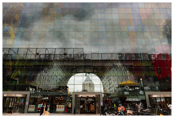 Blick auf eine monumentale, flache Glasfront, die oben den Himmel und Umgebung spiegelt. Im unteren Teil sieht man etwas durch das Glas hindurch. Man sieht in eine gewölbte Halle und durch die Halle hindurch ins Freie. Hinter der Halle sieht man einen Turm. Im Vordergrund gehen einige Passanten.