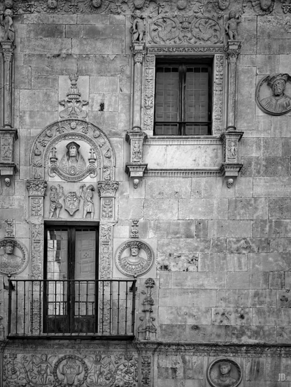 La imagen muestra una fachada de piedra ornamentada con relieves y esculturas en estilo renacentista. Se observan dos ventanas, una con balcón de hierro forjado. Alrededor de las ventanas hay medallones con rostros tallados, figuras decorativas y motivos florales y heráldicos que adornan la pared. La composición es simétrica y rica en detalles artísticos.