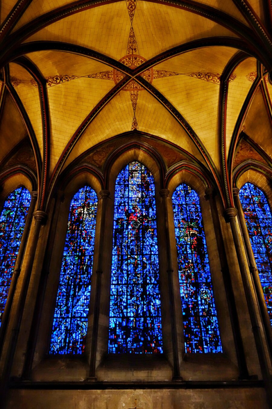 Interior of Salisbury Cathedral showcasing three towering lancet stained glass windows with intricate medieval designs in vibrant blues, deep reds, and golden yellows. The ribbed vaulted ceiling features intersecting pointed Gothic arches meeting at ornate bosses with intricate carvings. Tall stone columns with decorative capitals frame the windows.
