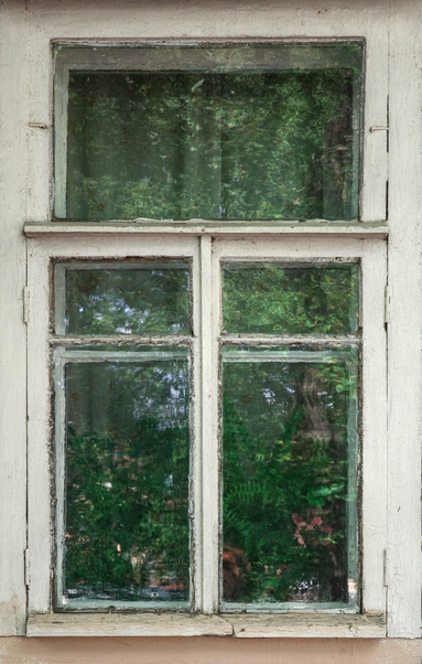 This color photograph captures a fragment of an old wooden window. The frame is covered in faded, peeling white paint that has cracked over time. The window is divided into three sections: a top horizontal transom and two vertical panes below. The glass is slightly cloudy, which gives the reflection a blurry, almost painterly character. A dense, lush green canopy of trees and bushes — likely growing just outside the window — is reflected in the glass. This reflection creates a "green glass" effect, completely filling the frame with vibrant spring greenery, contrasting with the white, peeling frame. The image conveys a sense of silence, coziness, and an old, comfortable house immersed in greenery.
