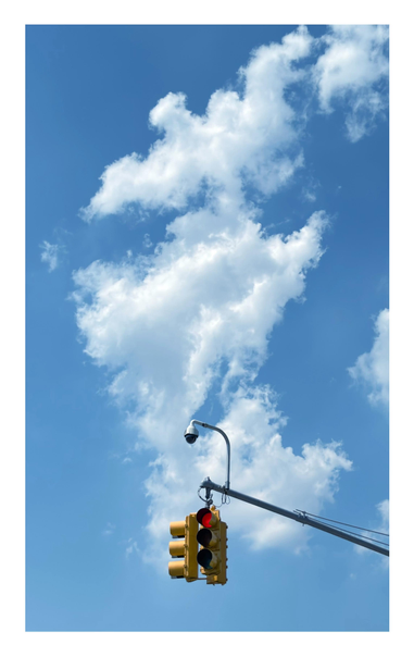 A traffic signal in yellow casing, with its red light activated, hangs from the end of a metal arm under a tall, vertical white cloud in a blue sky. Also above the traffic signal, sticking out from the arm, is what looks like a miniature streetlight; this is an EVP (Emergency Vehicle Preemption) sensor, which detects infrared signals from approaching emergency vehicles and overrides the normal sequence of traffic lights to allow the vehicle to pass through.