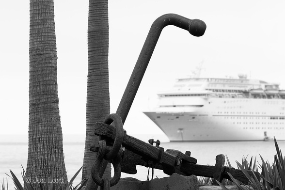 This is a black and white photo in landscape format of a large, old and corroded anchor lying on the waterfront in the foreground and an anchored cruise ship framed by the stock (Arm) of the anchor. Santa Catalina, California (2014).

In the foreground at the base of the image are the leaves of numerous arid climate plants, the slim and pointed leaves angled upwards at the sky. In the left half of the photo are the trunks of two large palm trees, the tops out of view beyond the upper edge of the frame. In the centre and extending away from the camera and to the right is our Admiralty Pattern Anchor, sometimes know as a 'Fisherman'. The sort of anchor seen in illustrations of old historic ships. The shackle end of the anchor is closest to the camera with the stock or arm angled upwards towards the sky, the last half metre bent downwards at ninety degrees with a ball shaped end. The shank stretches away from the camera with the two flukes (blades that dig into the sea bed) just visible lying on the ground. The light grey, almost silver, calm sea rises from the base of the image to a little less than a quarter of the way up in the photo. The horizon where the sea meets the sky is blurred and not clear. Framed by the arm of the anchor is the bow of an anchored cruise ship. The bow is in the centre of the photo with the ship viewed obliquely: the ship anchored diagonally across the photo with the bow and half the port (left side) in view and with the ship occupying half the width of the photo (the aft section of the vessel is out of view beyond the right edge). The ship is all white, partially blurred out of focus and has a small shore boat moored by an open hatch near the waterline. Beyond the ship is obscured by a thick summer morning's haze.