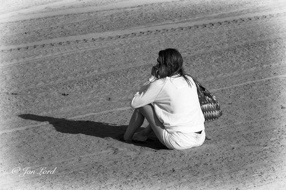 This is a black and white street photo in landscape format of a woman sitting alone on a beach and looking out to sea. Hermosa Beach, California (2012).

The entire image is filled with an expanse of smooth sand. Towards the top of the photo are numerous tyre tracks stretching across the image from left to right. Sitting very slightly to the right of centre is our lady, brightly lit from behind by the still low morning sun, her long shadow extends outwards towards the left edge of the frame. She is viewed obliquely with her back and left side in view and with her filling half the height of the photo. She has long dark hair, is wearing sun glasses, a white, long sleeved top and white shorts. She also has a large patterned bag over her right shoulder. Her knees are bent upwards in an inverted V shape with her arms resting on her knees. Her left hand is held up to her face, while her head is facing slightly to the left. 

Our candid subject, as I remember, is a young mother watching her son or daughter surfing in the Pacific Ocean.