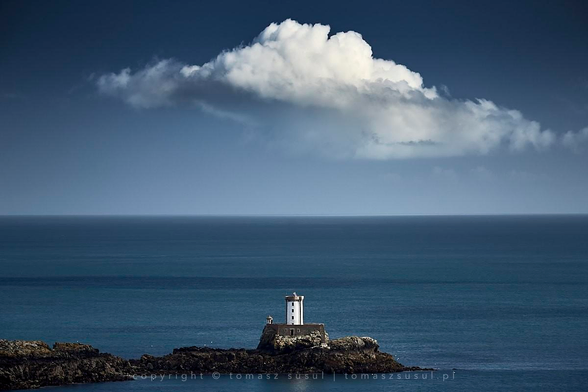 A compact white lighthouse stands perched upon a rugged rock formation amidst the sea. The surrounding water stretches toward the horizon, blending into deep blue tones of the sky in the distance. Above it, a massive, sculpted cloud towers over the sea, its immense size dwarfing the small lighthouse below. 