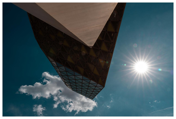 Wir stehen unter einem futuristischen und kantigen Gebäude und blicken direkt nach oben. Wir sehen das überhängende Gebäude (Beton und viel verspiegeltes Glas) und einen Sonnenstern.