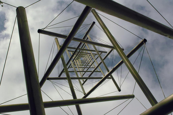 Blick von der Basis senkrecht nach oben in die aus Stahlstangen und Stahlseilen errichtete Turmskulptur. Die Stahlstangen sind durch Stahlseile miteinander auf Abstand verbunden. Das ergibt einen filigranen Turm.