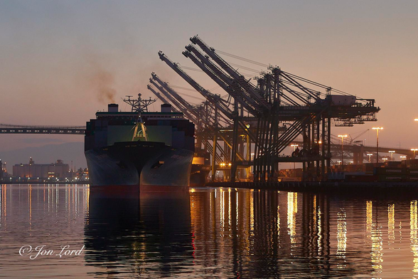 This is a colour photo in landscape format of a container ship leaving port at sunrise with numerous, raised gantry cranes to one side and a hint of the San Gabriel Mountains in the background. Port Of Los Angeles, California (2018).

The lower third of the photo is filled with a calm and gently rippled water with multiple reflections on the surface from the numerous port flood lights. In the centre is a recently departed container ship, only a few metres from the dock. The ship is viewed from the bow, 'head on' with neither side in view. The width of ship filling a quarter of the width of the photo, while the height of the ship fills about half the height of the image and is viewed from fifty metres away. The vessel is partially in shadow, the sun has yet to rise and is still just behind the mountain range in the background. The ship is riding high in the water with about two metres of red antifouling in view. Above the hull is dark green, Above the bow is a white floodlit mast, about four metres tall. Aft is a large metal screen or barrier protecting the container stack behind from breaking waves in a heavy sea. The stacked containers, deep in shadow, can just be made out behind the screen with the wheelhouse or bridge is visible above the container stack and two thirds of the way aft from the bow. The many horizontal, rectangular bridge windows nearly extend the entire width of the ship with the sky visible behind the ship through the windows on the sides. Behind the bridge are two short square funnels, the funnel on the left (camera's left) emitting a brown to orange wisp of smoke. Between the funnels is a frame supporting various radio and radar antennae. To the right of the ship is the container terminal with many neat rows of stacked shipping containers a short distance from the water. On the water's edge are seven giant gantry cranes, their booms all raised upwards at an angle of thirty or forty degrees. Behind our departing ship, a small portion of a moored further container ship can be seen. A short distance, about one km, behind this scene and spanning high above the water is the deck of a suspension bridge, though neither tower is view. In the far distance, about fifty km away, is the ridge of a mountain range. Above is a clear and cloudless sky, a golden orange lower down, especially on the right side of the photo, and graduating to a pale blue at the top of the image. 