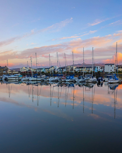 White sailboats and yachts are moored in a calm harbour at dusk. The still water holds a mirror reflection of the boats and soft blue and pastel pink sunset sky. In the background, townhouses sit at the edge of the harbour with distant mountains silhouetted on the horizon.