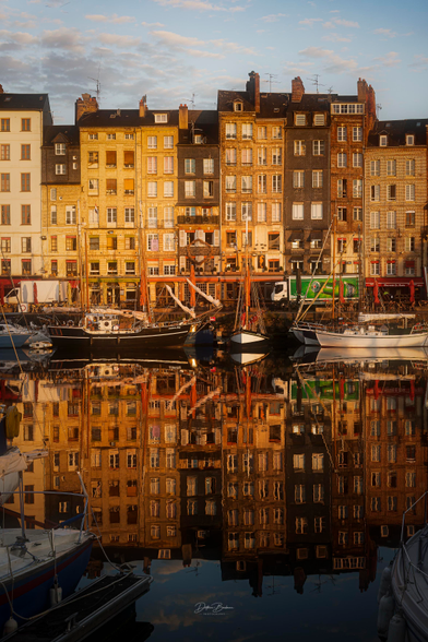 Eine malerische Hafenszene mit farbenfrohen historischen Gebäuden, die sich im ruhigen Wasser spiegeln. Im Vordergrund liegen verschiedene Boote vor Anker, und am Himmel sind sanfte Wolken zu sehen, die von goldenem Licht beleuchtet werden.