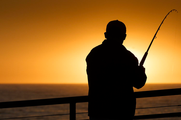 This is a colour photo in landscape format of the silhouette of the back of a sports fisherman, standing on a pier and fishing in the sea with the setting sun in front of him. Redondo Beach, California (2014).

Sloping downwards gently from the lower right side towards the lower left corner is the metal railing at the edge of the pier, between the rails are some cables stretched parallel to the poles for added protection. The sea rises from the base of the photo upwards to the quarter up level where there is a crisply defined horizon. The sea water is blurred out of focus but moderate sized waves are visible. Standing slightly to the right of centre is our angler (sports fisherman). His back is to the camera, viewed from the waist upwards, with his height occupying most the height of the image and viewed from a few metres away. He is holding his fishing rod (fishing pole) in his right hand with the rod extending diagonally upwards into the upper right corner of the photo. The far end of the rod is bent downwards, so perhaps he has caught a fish? The setting sun is immediately in front of him with his body obscuring all of the sun, leaving a bright glow or aura surrounding our fisherman. The remainder of the clear and cloudless sky is a golden to orange luminous glow: partly from the sun caressing the horizon, partly from the Southern California's dubious air quality and partly because I didn't correct the colour balance of the photo. The golden glow is reflected over the entire image. 

