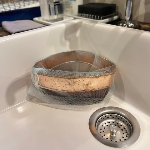 Ein dreieckiges Spülbecken-Sieb aus Kupfer, überzogen mit einem feinmaschigen biegsamen Metallvlies in der Ecke eines weißen Spülbeckens, zum Aufnehmen kleinerer Speisereste.