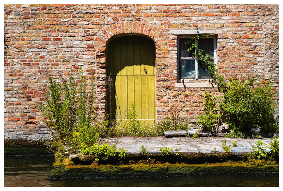 Blick auf ein Backsteingebäude. Man sieht eine grüne Tür und ein Sprossenfenster daneben. Auf einem kleinen Vorplatz wuchern Pflanzen. Davor ist ein Wasserlauf zu sehen. Vermutlich kann die Tür also nur per Boot erreicht werden.