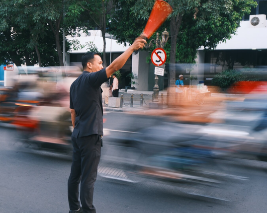 Security man waving a traffic baton prepares to stop vehicles on the road as bikes wheezes past him
