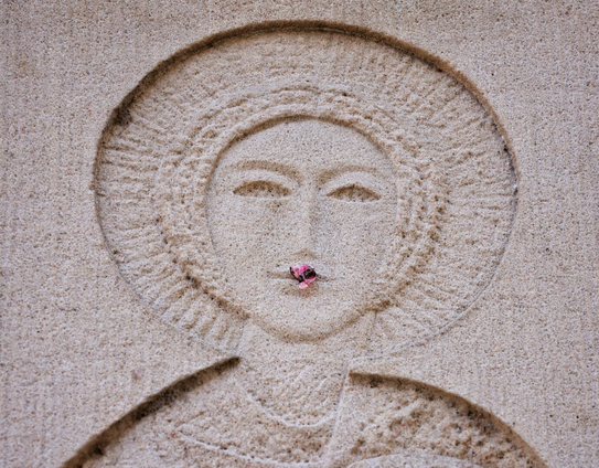 Close-up of a stone carving depicting Saint Dorothea’s face, surrounded by a circular halo. A small pink flower is stuck to her lower lip, giving the illusion of lipstick. The rough, textured stone and the saint’s serene, stylised features are clearly visible.