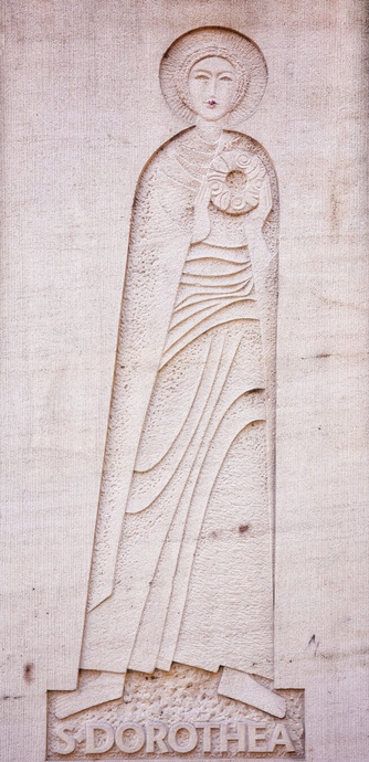 Full-length stone relief of Saint Dorothea, wearing flowing robes and a halo. A pink flower is affixed to her lip. The base features the inscription S DOROTHEA in capital letters. The carving shows fine details of her drapery, posture, and the halo’s concentric lines.