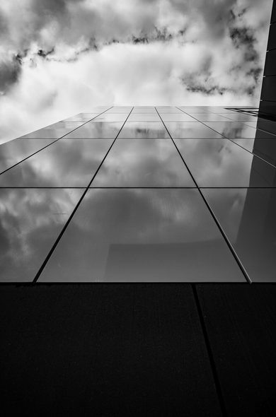 The image is a black and white, low-angle shot looking upwards at a modern building. The dominant elements are a dark, textured base and a vast expanse of glass panels reflecting the sky. The composition is stark and geometric, with strong diagonal lines formed by the edge of the building and the vertical and horizontal lines of the glass panels. The sky, filled with dramatic, dark clouds, occupies the upper portion of the frame, its reflection creating abstract patterns on the glass.