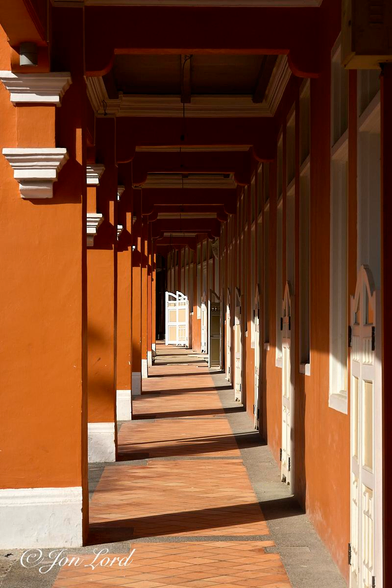 This is a colour street photo in portrait format of a deserted, saffron coloured, covered walkway forming the street-side pavement in a restored, historic colonial-era district. Keong Saik, Singapore (2012).

Stretching away from the camera is the covered walkway. Its extends for about ten metres before turning to the left and out of view. The camera is about a metre and a half above the ground, positioned in the centre of the one and a half metre wide walkway and aligned towards the end of the passageway. The floor is made from orange coloured terracotta tiles laid in a diamond pattern with grey stones lining the edges, the passageway slopes upwards ever so slightly with increasing distance. On the left side is a row of seven square pillars that support the upper floor of the street side of the building. Each has a white base supporting the masonry pillar, painted a rich, vibrant, saffron (orange to brown colour). On the right side is the masonry wall of the building, also painted that attractive saffron colour. Each shop, business or house has a white painted wooden door with two arched shutters rising from the floor upwards a metre and a half high and opening outwards. Either side of the door are tall, vertical rectangular windows that reach upwards to the ceiling of the walkway. This pattern of window, door, window repeats along the corridor and out of view as it bends to the left in the distance. The ceilings are equally ornate with a wooden lintel crossing the walkway at right angles in the gap between the two windows. These lintels make up a square with a white painted surround that repeats along the corridor. 

