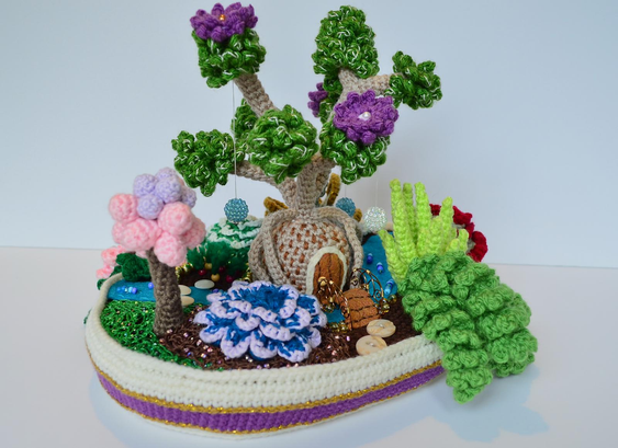 Full view of the fairy garden from the front. In the center is a small tree with a lumpy base and some branches resembling broccoli florets with purple flowers. Three large beads are hanging from the branches. The base of the tree has a felt door in it. There's a felt stream running from one side to the other. A bridge crosses the stream right in front of the door. Behind the tree house there is a turnip garden. In front of the tree house there are a few smaller decorative plants: one that looks like cotton candy, one rosette, one fluffy trailing plant, and one pointy aloe. The entire scene sits in a wide oval planter. Everything is crocheted, except for a few embellishments in felt, beads/buttons, and wire.
