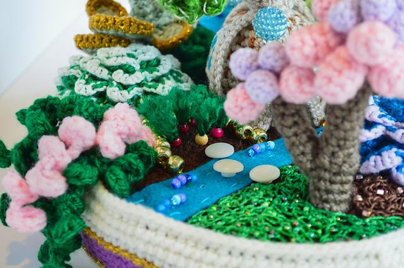 Closeup of the mini turnip garden. The turnips are made of small beads with loose green yarn plies sticking out the top. The garden plot is fenced off with gold pony beads. The turnip garden is next to a felt stream, which has three stepping stones made of buttons.