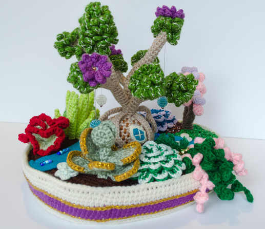 Fairy garden back view. The back of the broccoli tree has a window made of felt. Different plants are visible from this angle. On the far side of the stream there's a wrinkly lettuce-y plant next to an aloe. On this side of the stream there's a silver dollar jade with sparkly gold edges, a rosette that could be an echeveria, and a trailing vine.