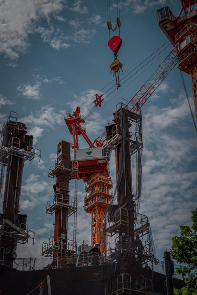 A dramatic low-angle view shows steel pillars reaching into a cloudy sky at a busy construction site. Bold red and orange cranes rise high above the rising metal frame. Small figures of workers navigate scaffolding, while a vibrant red crane hook hangs overhead, capturing the powerful energy of building a new horizon.