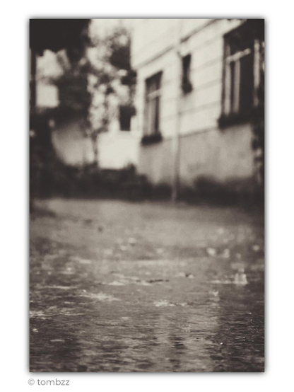 Analog black-and-white photograph in a vintage style, taken in the rain from a very low perspective. The foreground is filled with wet pavement, where falling raindrops, small splashes and circles spreading across a thin layer of water are visible. This is where the image holds slightly more focus, emphasizing the texture of the puddles and the shimmering reflections of light. In the softly blurred background stands an old, classically styled building with a pale façade divided by horizontal bands. Dark rectangular windows, some with small sills or decorative frames, create a quiet rhythm across the wall. On the left, indistinct silhouettes of trees, shrubs and part of a neighbouring building can be seen. The softness, grain and muted shades of grey give the photograph a nostalgic, slightly dreamlike atmosphere, like a rainy day preserved on an old strip of film.