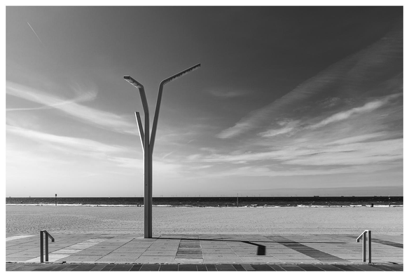 Moderne Y-förmige Straßenlaterne an einer leeren Strandpromenade. Dahinter liegen Sandstrand, Meer und ein weiter Himmel mit langgezogenen Wolken; am Horizont sind Windräder und Schiffe zu sehen.
