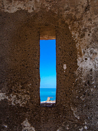 A narrow, rectangular opening in a thick, textured stone fortress wall frames a bright blue sky fading into a calm turquoise sea, with a small port structure visible at the bottom.