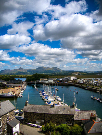 A scenic view looking over Porthmadog Harbour to the mountains of Eryri (Snowdonia) in the distance under a bright blue sky filled with white fluffy clouds.
In the foreground, stone buildings sit on the edge of the harbour which is filled with sailboats and yachts. Multicolored contemporary townhouses line the right side of the water. In the distance, the jagged peak of Cnicht can be seen, aside those of Moelwyn Mawr and Moelwyn Bach.