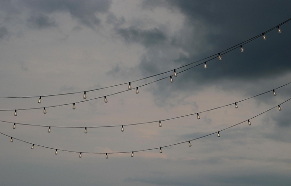 Photograph of four strings of lights, bulbs lit and crossing the image from left to right, while behind the sky is cloudy and threatening at sunset, with very dark clouds and some pinkish hues near the horizon.

Photographie de quatre guirlandes de lumières, les ampoules allumées et traversant l'image de gauche à droite, alors que derrière le ciel est nuageux et menaçant au coucher du soleil, avec des nuages très foncés et quelques teintes rosées près de l'horizon.