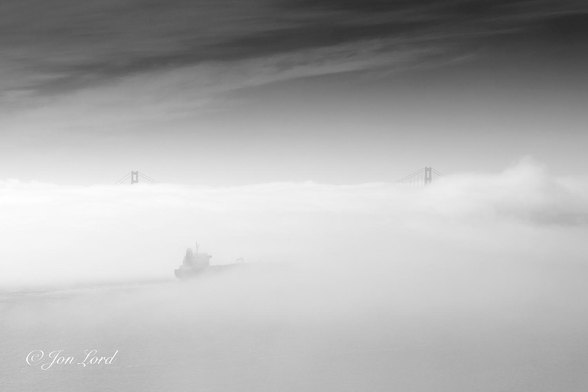 This is a black and white photo in landscape format of a cargo ship passing through a bank of thick sea fog and approaching the Golden Gate Bridge. San Francisco Bay (2017).

A mass of sea fog fills the lower half of the photo, a little thinner in the lower left corner, a little thicker and more dense to the right. To the left and below centre in the zone of thinner fog, is the partial view of a cargo ship, with just the stern's white superstructure and less than a third of the aft section of the black hull in sight. The ship is underway from left to right, the stern and starboard (right) side in view and with what is visible of the ship, filling a little less than a quarter of the width of the photo. Aft of the ship is a long and narrow gap in the fog marking the vessel's passage. Either side of the photo's centre and about one km ahead of the ship are the tops of two towers of a large suspension bridge with only the upper ten metres and some of the suspender cables in view. Above is a nearly clear sky, rendered dark grey with a gossamer thin, wisp of high cirrus clouds, mainly in the upper left side of the image.
