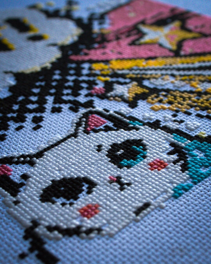 An extreme close-up of a cross-stitch project in progress. In the foreground, a cute anime-style white cat with large blue eyes (only one filled in) and pink cheeks. Above the cat, sections of a pattern with shooting stars in black, pink, and yellow are completed, while unstitched areas of the fabric are visible between the stitched areas, showing the piece is unfinished.