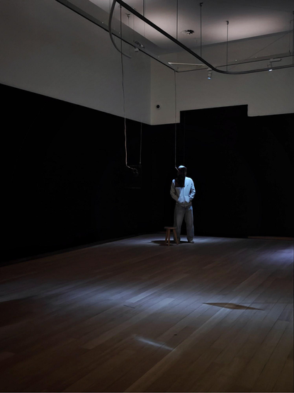A photo of a dimly lit room with a wooden floor. A person in a hoodie stands near a small object suspended from the ceiling.