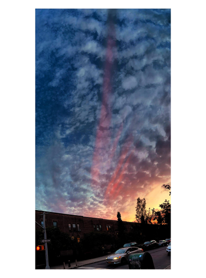 Photo of a residential street at sunset with a red rainbow in the wide sky overhead. The sun sinks behind trees in the distance at right, casting a golden glow on that corner of the sky, and throwing the neighborhood — low-rise apartments, parked cars — into shadow. The rest of the sky is a deep evening blue with white clouds all across it. Bands of red light stretch overhead — really a bow, but so large that the photo only shows a nearly vertical portion of their arc.