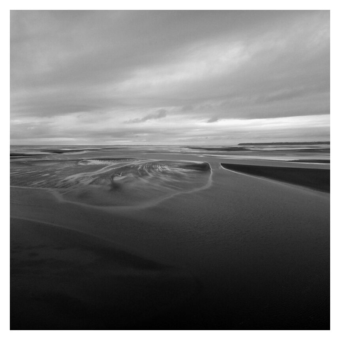 Vue de haut, au format carré, en noir et blanc, une vaste baie, où la mer, les bancs de sables et les nuages dessinent des formes abstraites.