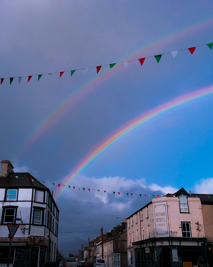 A bright double rainbow arcs across the sky over Stryd Fawr (High Street), Porthmadog. The secondary rainbow above lies in a darker patch of sky and shows reversed colours from the original rainbow which is has a bright blue sky background. On the right, a cream-colored building with a tiled sign reading "Richard Newell Limited." To the left is a white building with timber framing. Decorative red, white, and green bunting (the colours of the Welsh flag) hangs overhead.