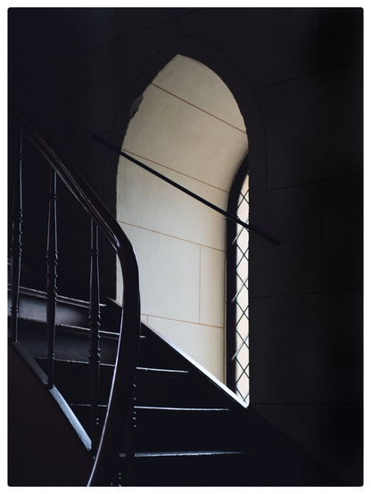 Monochrome interior image of a window set into a gothic arch with light shining on a wooden staircase with carved banisters curving towards the viewer.