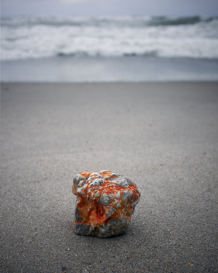 The photo shows a cubic rock or stone at a sandy beach with a wavy Mediterranean Sea in the background. The rock or stone has interesting orange and red parts between the grey.